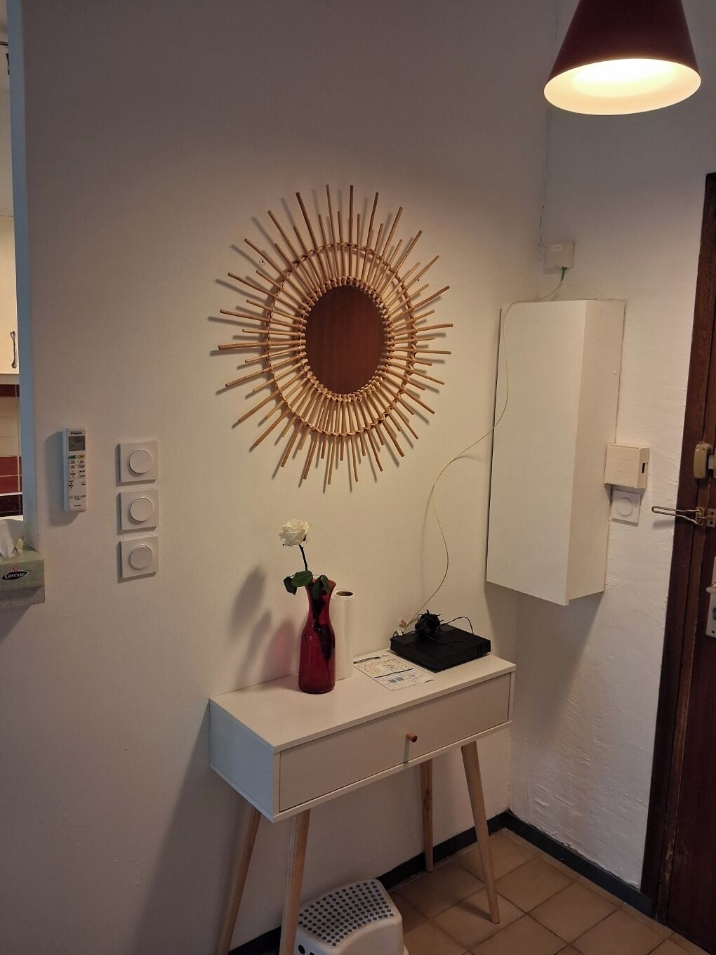 Una habitación con un espejo tipo sol montado en la pared, un escritorio blanco con un florero, un armario blanco y un calentador portátil.