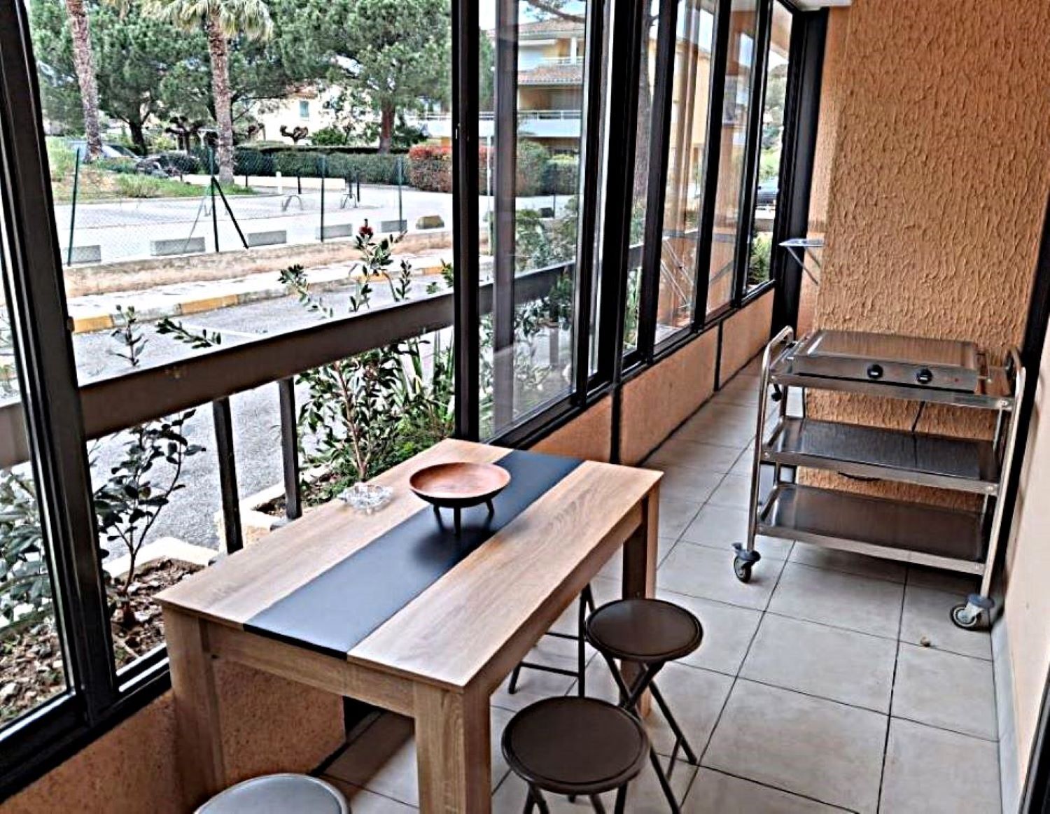 A modern interior with a wooden table and stools, large windows offering a view of the outdoors, and a metal rack on the right.