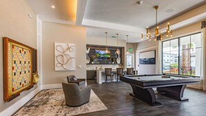 A modern living room with a pool table, comfortable seating, a kitchen area, and large windows offering a view. - Pinnacle Apartments W Amenties by Heavenly Homes (Orlando)