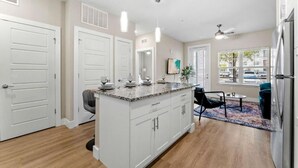A modern kitchen with white cabinets, a granite countertop, and a central island. There are two pendant lights, a ceiling fan, and a window with blinds. - Pinnacle Apartments W Amenties by Heavenly Homes (Orlando)