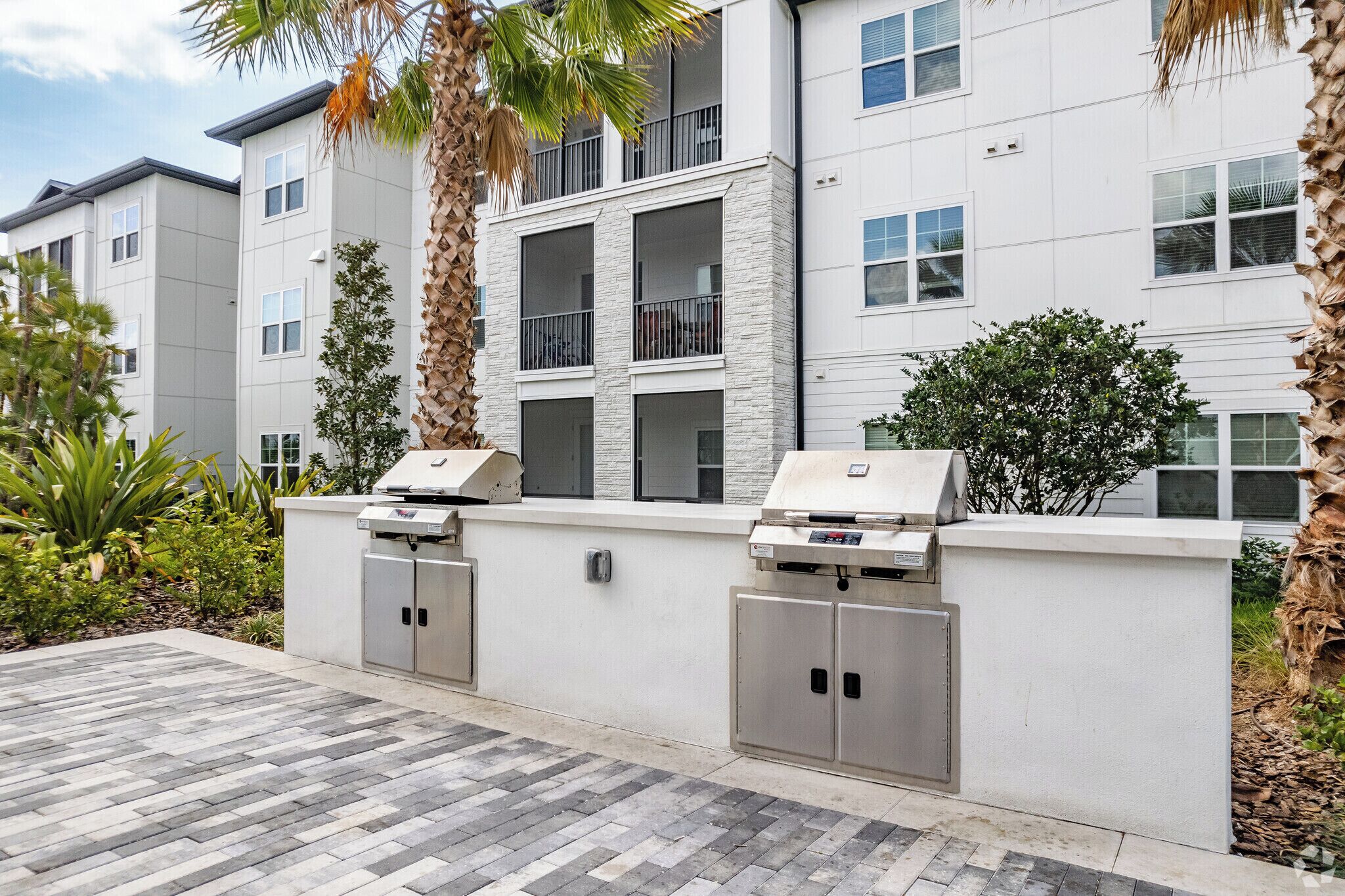 A modern apartment building with outdoor grilling stations, a paved patio, and a landscaped garden.
