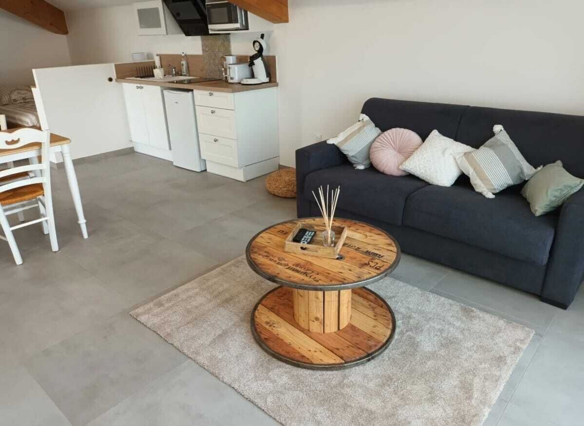 A modern living room with a round coffee table, a sofa with cushions, a kitchen area with white cabinets, and a dining table with chairs.