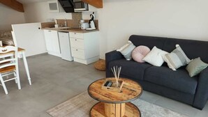A modern living room with a round coffee table, a sofa with cushions, a kitchen area with white cabinets, and a dining table with chairs. - Charming studio-cabin in quiet location near Nîmes and beaches (Bouillargues)