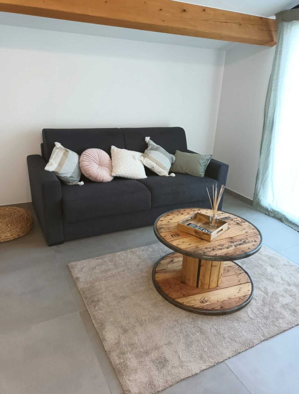 Un salon avec un canapé foncé, de nombreux coussins, une table basse ronde en bois sur laquelle se trouvent un plateau et des bâtonnets, et un panier en osier.