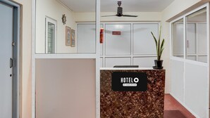 Ein Hotelrezeptionstresen mit einem Schild, auf dem 'HOTELO Company Serviced' steht, ein Deckenventilator, ein Topfpflanze und eine Tür links.