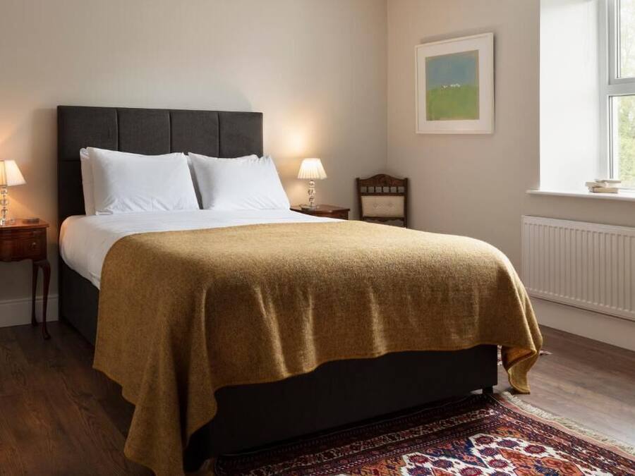 A bed with a dark headboard, two bedside tables with lamps, a framed picture on the wall, and a window with a view of trees.