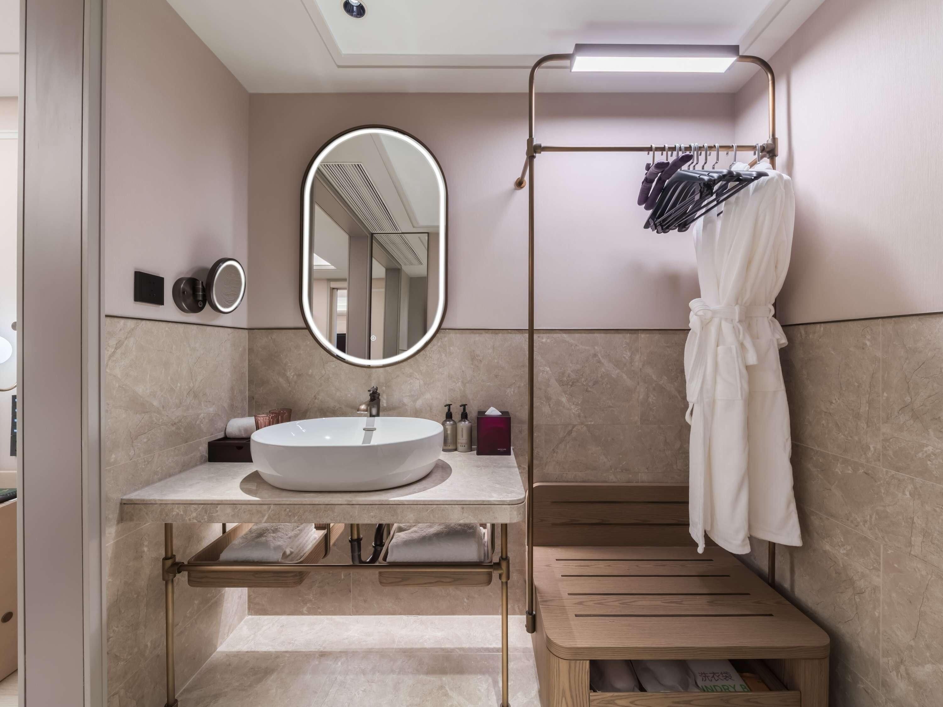 A modern bathroom with a large oval mirror, a round sink, and a wooden bench.