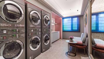A laundry room with four commercial washers and dryers, a small table with a vase, and a seating area with a sofa and armchairs.