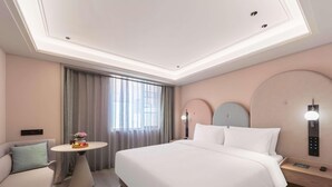 Quarto de hotel com uma cama grande, mesa de cabeceira, pequena mesa redonda com uma cesta de frutas, sofá e janela com cortinas.