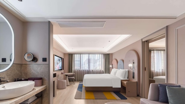 Quarto de hotel moderno com uma cama grande, uma mesa redonda, um sofá, um banheiro com pia e espelho, e uma televisão.