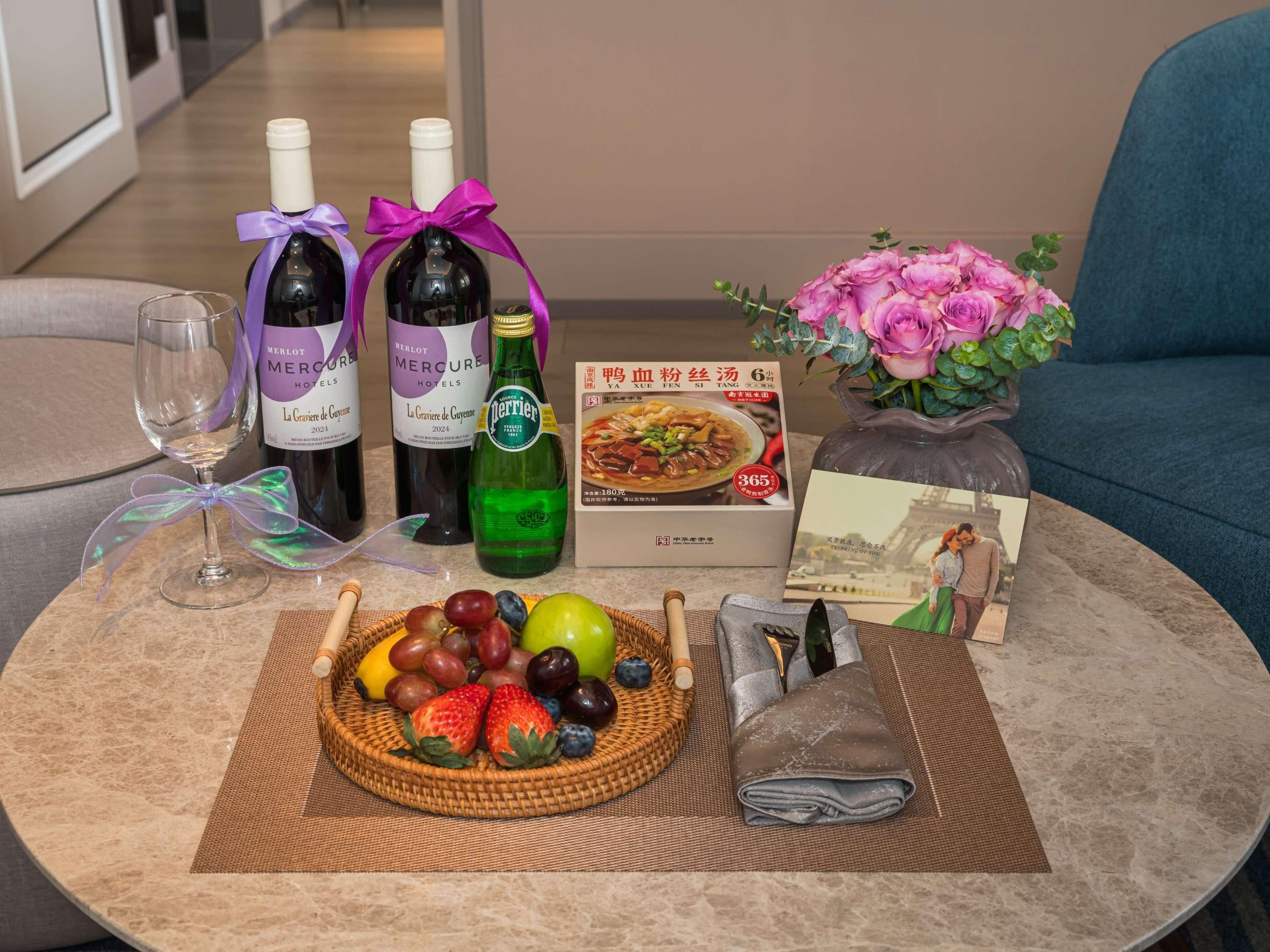 A table with two bottles of wine, a box of food, a bouquet of flowers, and a basket of fruit.