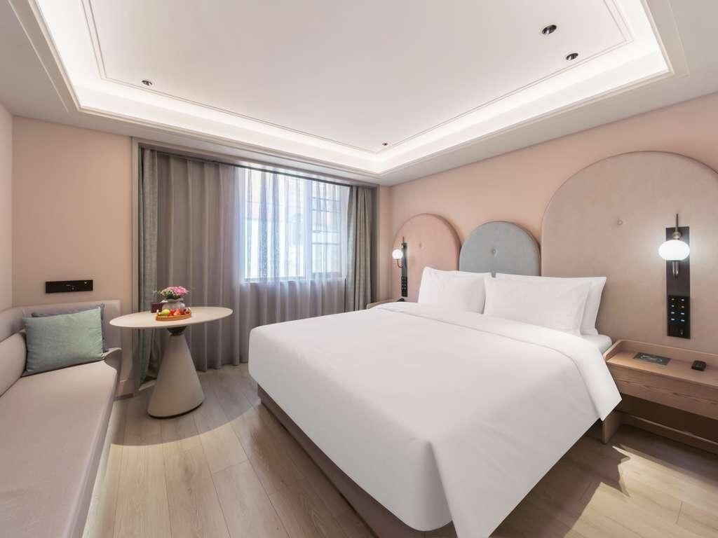 A hotel room with a large bed, a round table with a fruit bowl, a sofa, and a wall-mounted control panel.