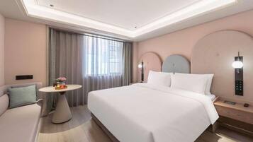 A hotel room with a large bed, a round table with a fruit bowl, a sofa, and a wall-mounted control panel.