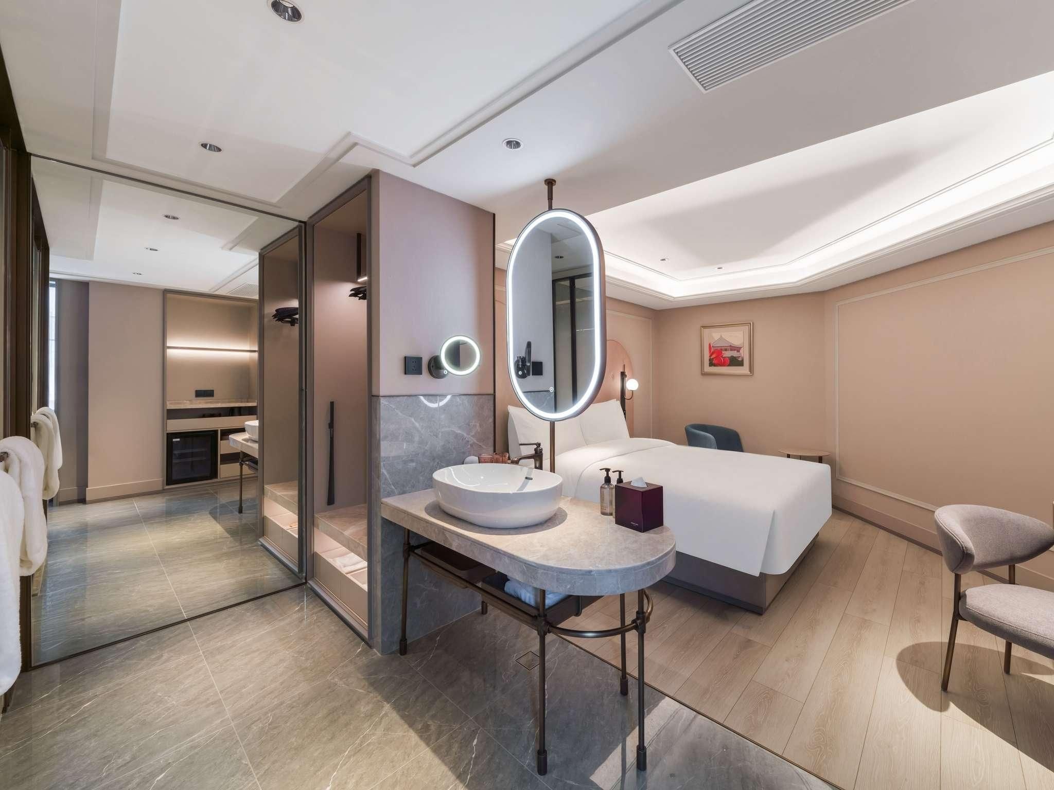 A modern hotel room with a round mirror, a bedside table, a bed, and a chair.