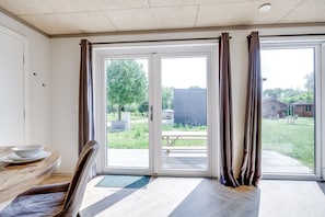 A room with a dining table, chairs, a bench, and a view through large sliding glass doors to an outdoor area with greenery and a building.