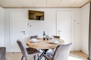 A dining area with a round wooden table, four grey upholstered chairs, a bottle of wine, and white plates. - Cube 96 | 4 persoons chalet in Zennewijnen (Zennewijnen)