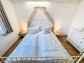 Un lit bien fait, avec une literie à motifs floraux et une tête de lit, deux lampes de chevet, une commode blanche et une fenêtre avec des rideaux.