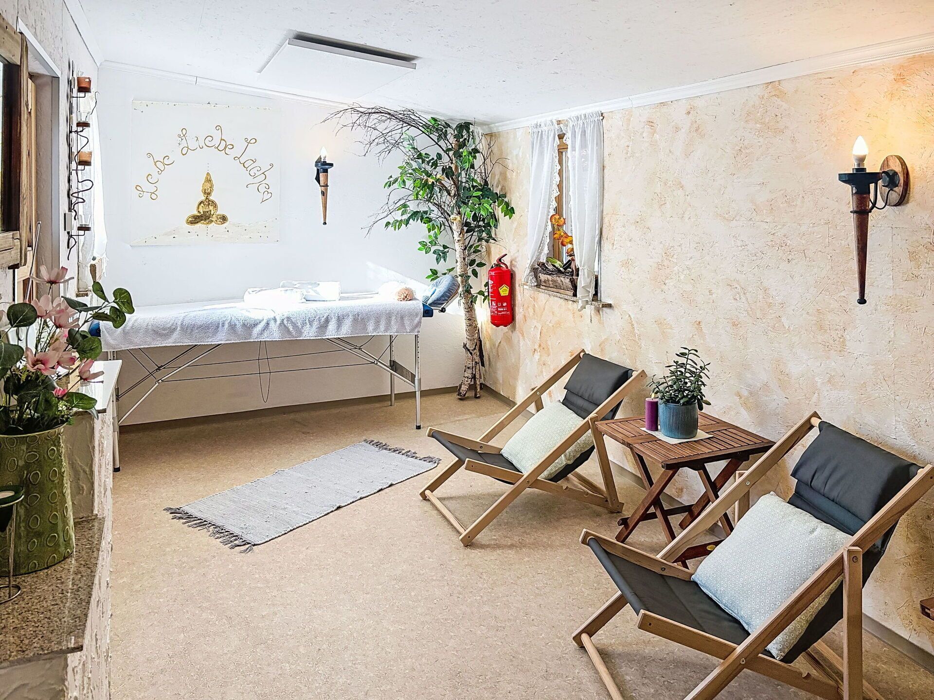 A room with a bed, two wooden chairs, a small table, a potted plant, a fire extinguisher, and a wall-mounted light fixture.