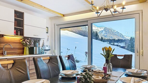 Un coin repas avec une table dressée pour quatre personnes, une cuisine équipée d’appareils modernes et une grande fenêtre offrant une vue sur les montagnes.