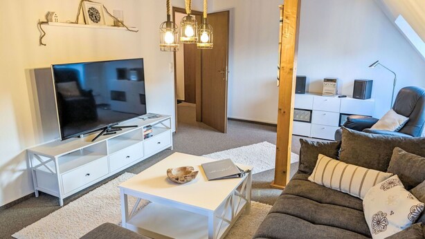 Una sala de estar moderna con televisor de pantalla plana, mobiliario blanco, techo con vigas de madera y un rincón de estar acogedor.