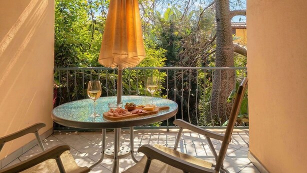 Une table ronde avec deux verres à vin et une assiette de nourriture, un parasol jaune et une vue sur des arbres et une clôture.