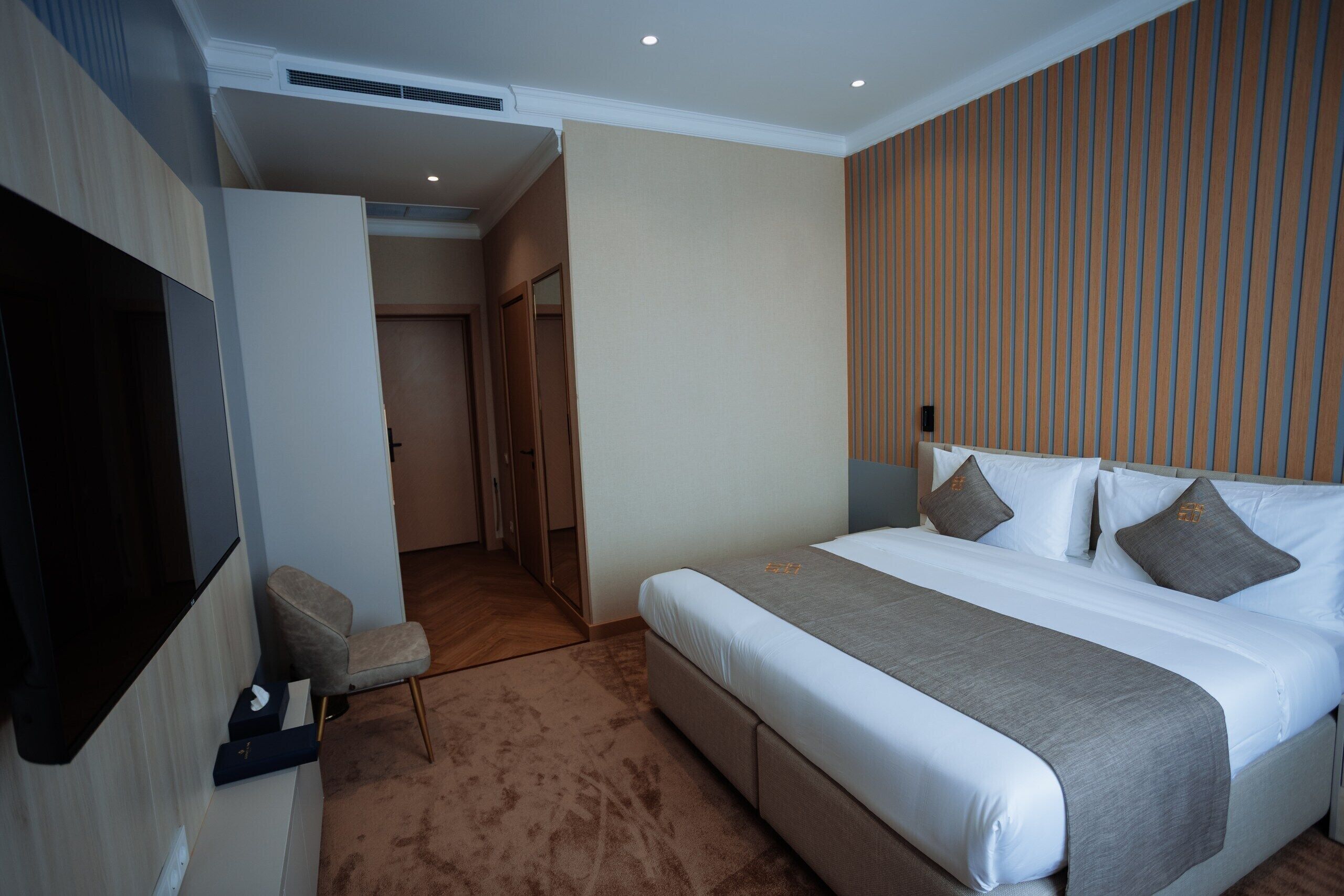 A hotel room with a bed, a TV, a seating area, and a door leading to a hallway.