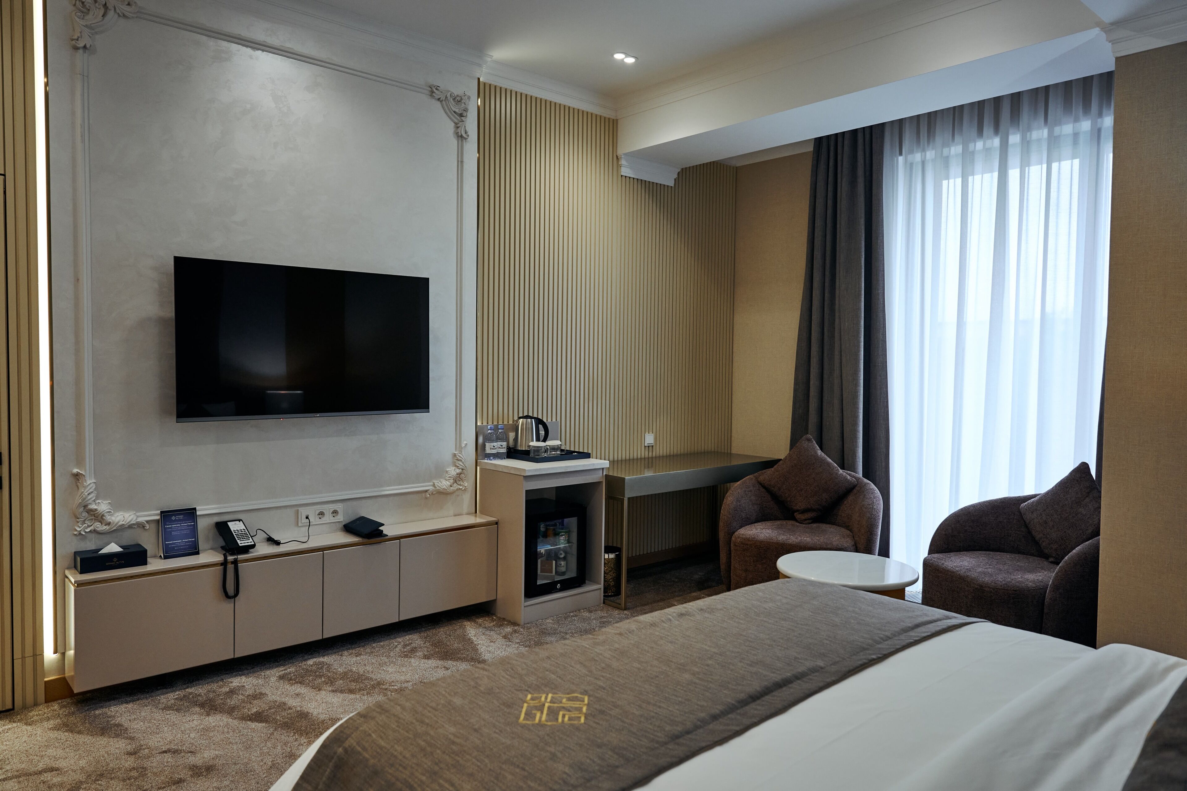 A hotel room with a large bed, a TV mounted on the wall, a desk with a computer, a chair, and a sofa.