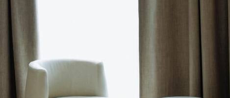 A modern room with a white armchair, a small round table, and a large window with curtains.