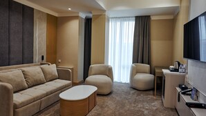 A modern hotel room with a sofa, armchairs, a coffee table, a TV, and a desk with a kettle.