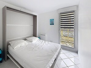 Ein Schlafzimmer mit Bett, Fenster mit Jalousien, einem kleinen Nachttisch und einer Wandleuchte.
