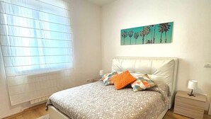 Une chambre à coucher comprenant un lit, une table de chevet, une lampe, une fenêtre avec des stores et une œuvre d’art murale.
