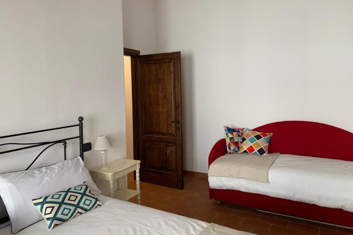 A bedroom with a bed, a red sofa, a wooden door, and a small bedside table.