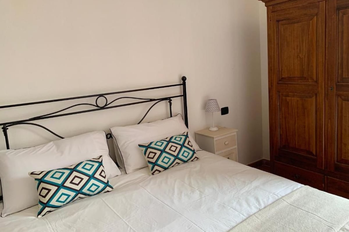 A bed with white bedding and patterned pillows, a bedside table with a lamp, and a wooden wardrobe.