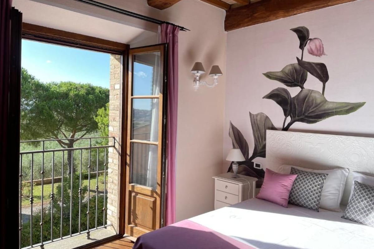 A bedroom with a large bed, a nightstand, a wall-mounted lamp, and a balcony with a view of trees.