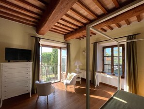 A room with a wooden ceiling, a white dresser, a chair, a small table with a lamp, and large windows with curtains. - Villa Preselle Country Resort (Scansano)