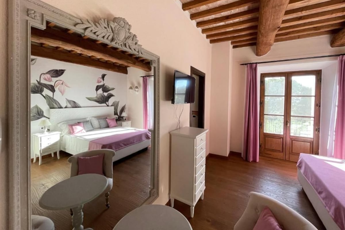 A bedroom with a bed, a dresser, a TV, a window with curtains, and a mirror reflecting the room.