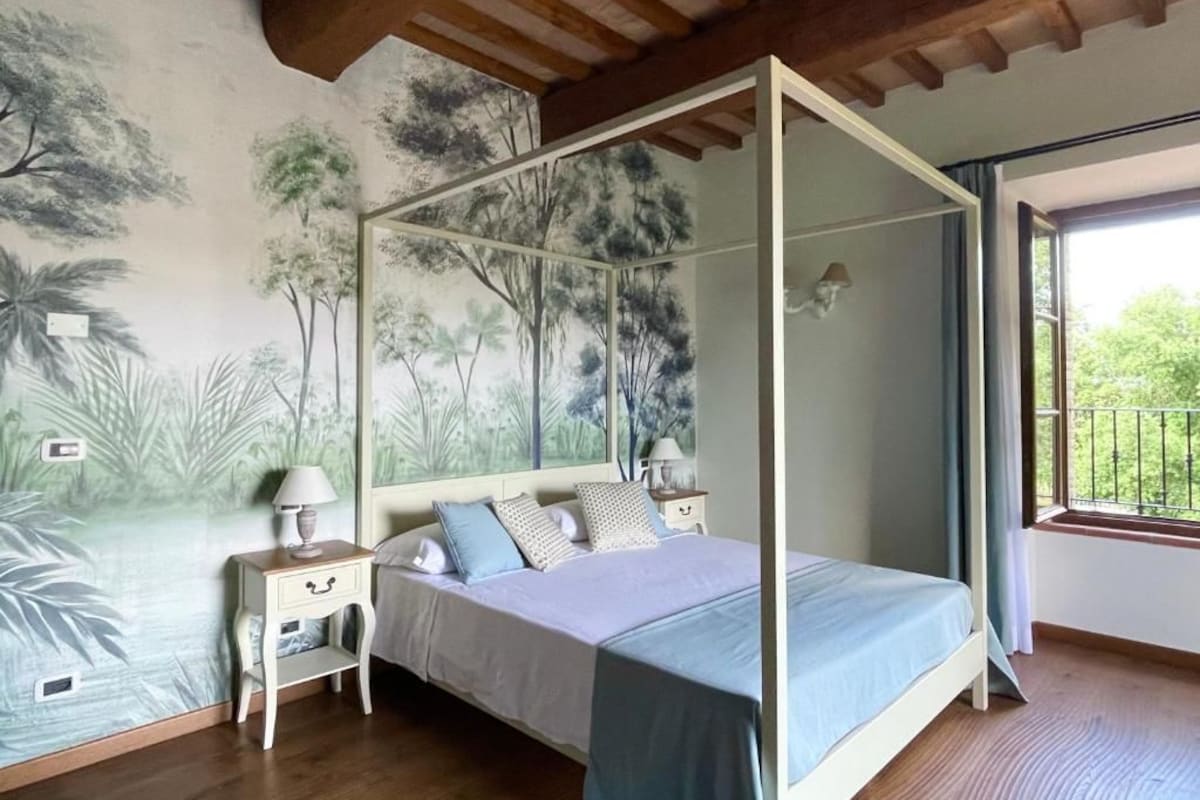 A bedroom with a four-poster bed, a nightstand with a lamp, and a mural of trees on the wall.