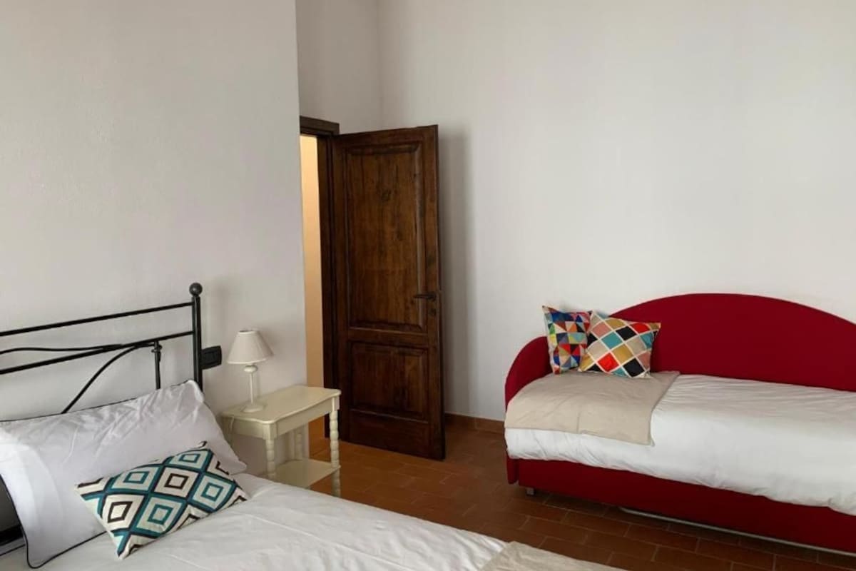 A room with a bed, a red sofa, a wooden door, and a small bedside table with a lamp.