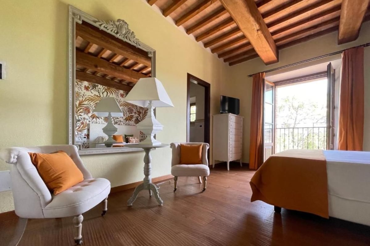A hotel room with a wooden ceiling, a bed, a sofa, a chair, a table with a lamp, and a view of the outdoors.