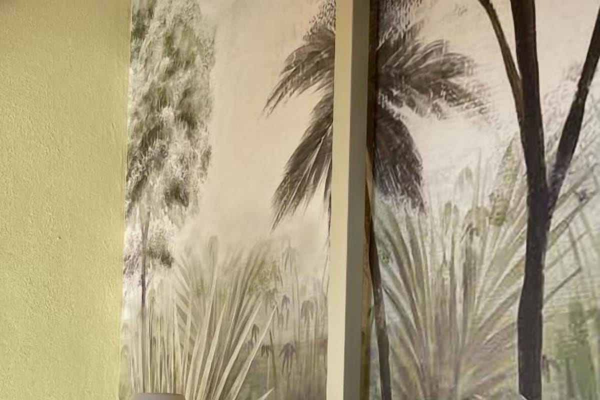 A bedroom with a bed, a nightstand with a lamp, and a wall mural of tropical plants.
