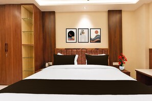 Une chambre d’hôtel avec un grand lit, une armoire en bois, une table de chevet, un vase avec des fleurs et trois tableaux encadrés au mur.