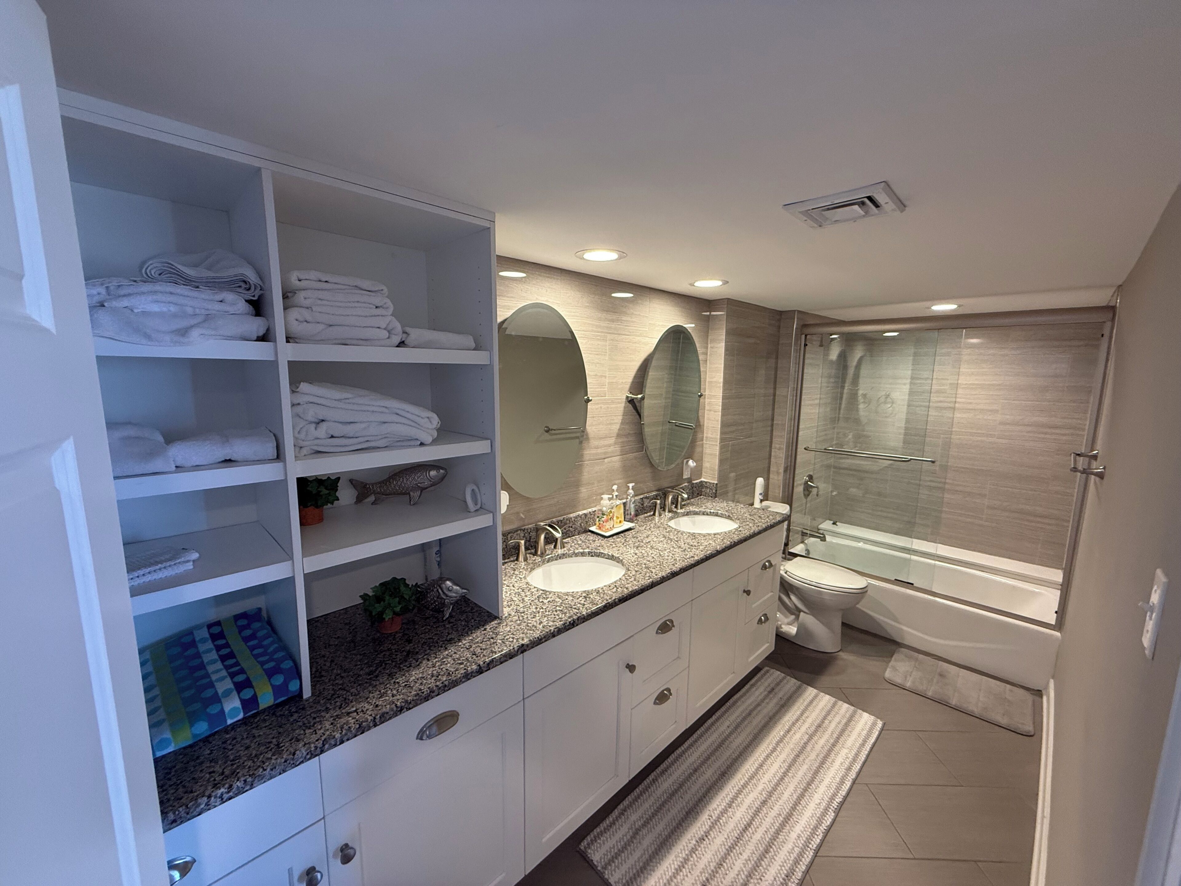 A modern bathroom with a walk-in shower, two sinks, a large mirror, and built-in shelves with folded towels.