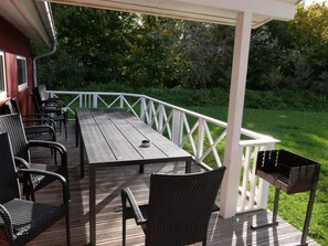 A covered outdoor deck with a wooden table and wicker chairs, a grill, and a white railing. - 4 Star Holiday Home in Grossenbrode (Grossenbrode)