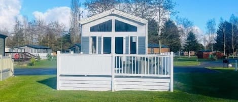 A small, single-story building with a white exterior and a balcony, surrounded by a grassy area and trees.