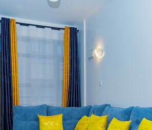 Habitació amb un sofà blau, coixinets grocs, una finestra amb cortines blaves i grogues, i un llum fixat a la paret.