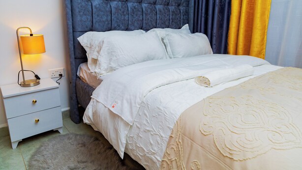Cama bem arrumada com roupa de cama branca, cabeceira azul e abajur.