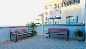 Um terraço no telhado com duas bancas de madeira, plantas em vasos e um edifício ao fundo.
