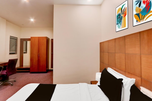 A hotel room with a bed, a desk, a chair, a wardrobe, and two abstract paintings on the wall.