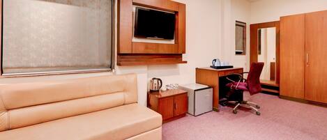 A room with a sofa, a TV mounted on the wall, a desk with a chair, a small refrigerator, and a wardrobe.