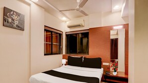 A hotel room with a bed, a ceiling fan, an air conditioner, a window, a mirror, and a bedside table with a lamp and a vase of flowers.